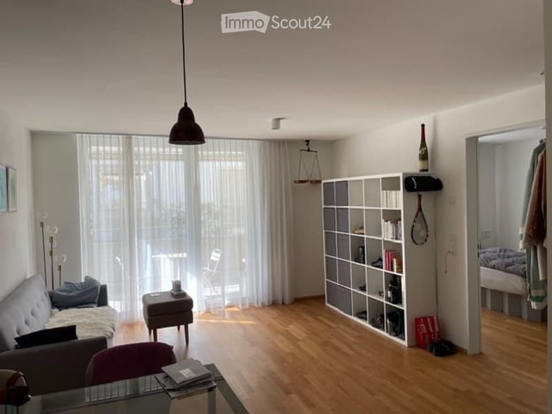 A modern, well-lit living room with wooden flooring, large windows covered by sheer curtains, a grey sofa with pillows, a small wooden stool, a white modular bookshelf, a glass coffee table, and a hanging pendant light.
