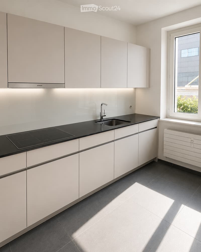 Modern kitchen with white cabinets, black countertop, induction cooktop, sink, and radiator. Natural light streams in from a window on the right.