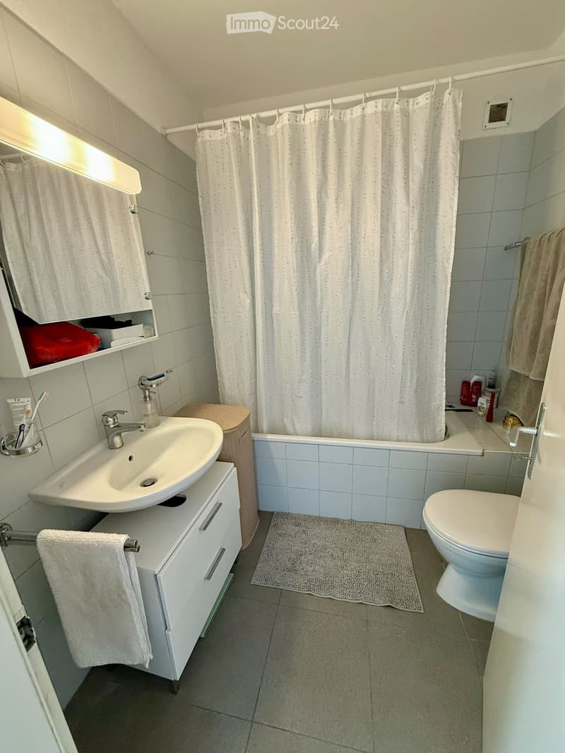Modern bathroom with white tiles, a sink with a modern faucet, a toilet, a bathtub with a shower curtain, a towel on a rack, and a small bathroom cabinet with a mirror above the sink.