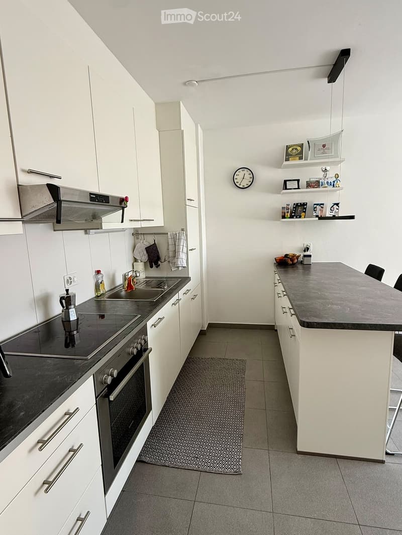 Modern kitchen with white cabinets, stainless steel appliances, island with countertop, sink, stove, and coffee maker. Shelves with decorative items and a clock on the wall.