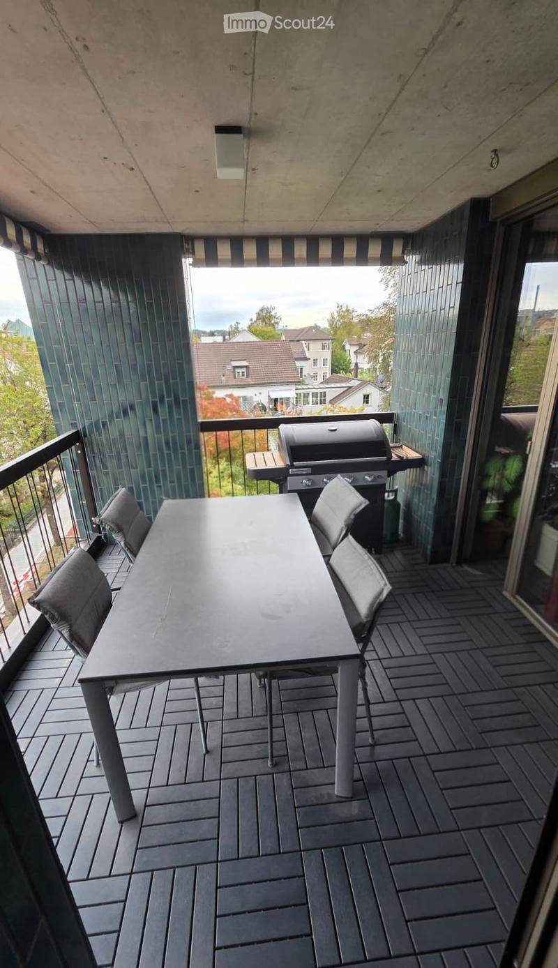 Balcony with a dark wooden deck, a rectangular table with four chairs, and a grill in the background, overlooking a residential area.