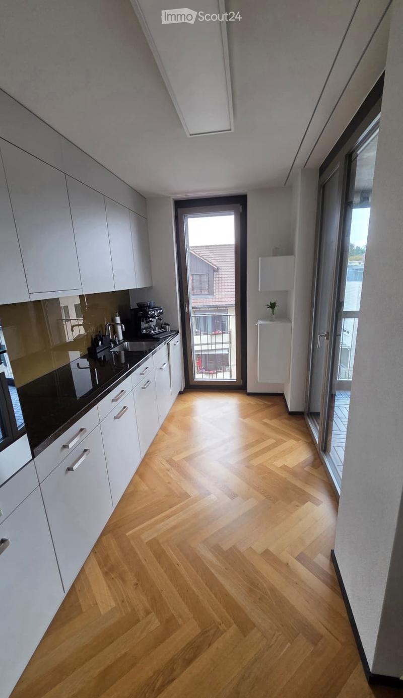 Modern kitchen with white cabinets, wooden herringbone flooring, and a coffee machine on the counter. A sliding glass door leads to a balcony.