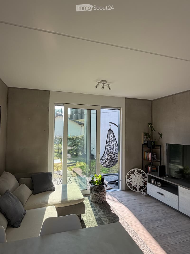 Modern living room with large sliding glass doors leading to a balcony, a grey sectional sofa, a round coffee table, a TV stand, and decorative items including a hanging chair and a wall-mounted decorative piece.