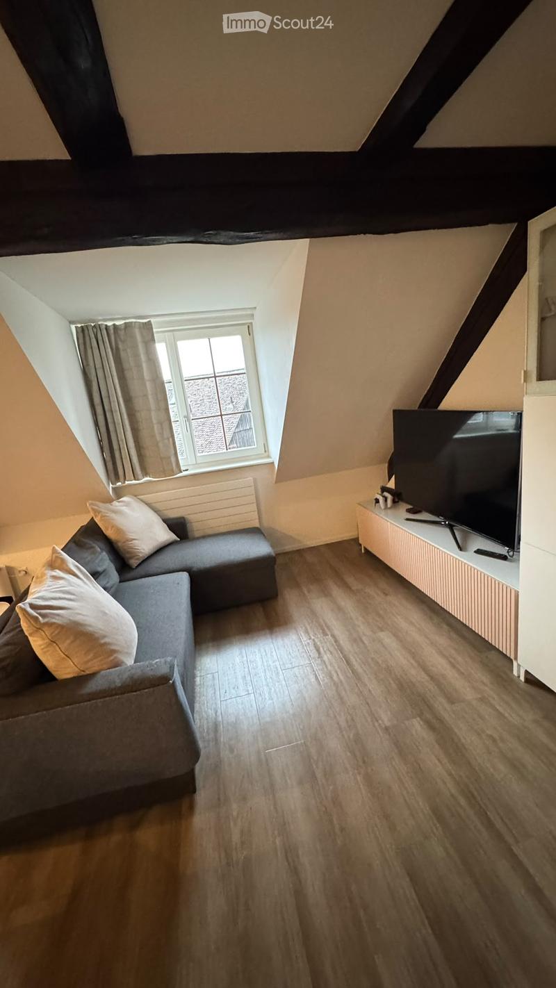 Cozy attic living room with wooden flooring, grey sectional sofa, television on a stand, and a window with curtains.