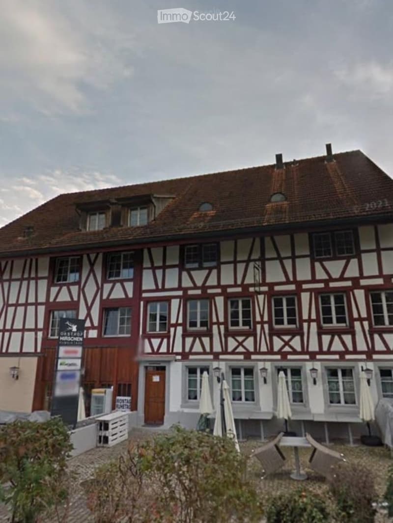 Traditional half-timbered building with red and white facade, multiple windows, and a brown tiled roof. Outdoor seating area with tables and chairs is visible in front.