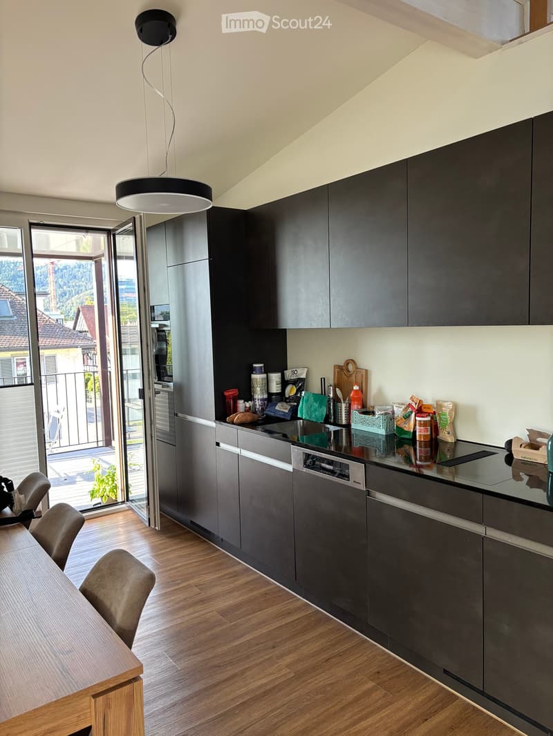 Modern kitchen with dark grey cabinets, stainless steel appliances, wooden flooring, and a pendant light. The kitchen features a dining area with a wooden table and chairs, and an open sliding door leading to a balcony with outdoor views.