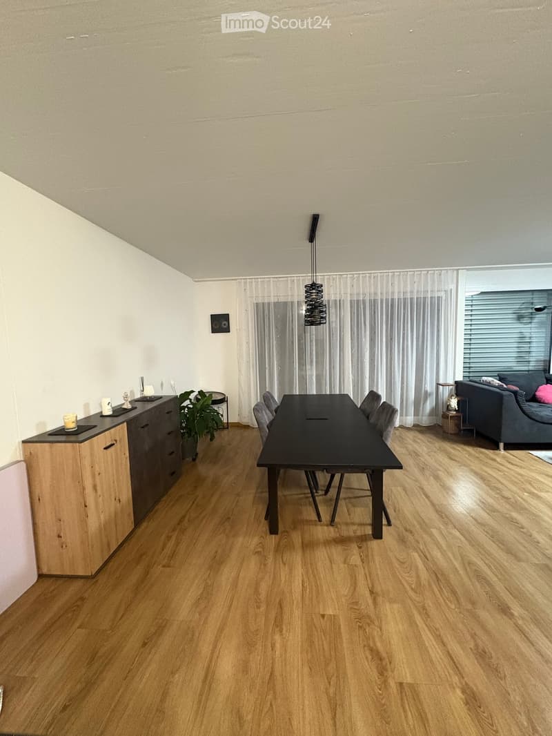 Modern dining and living area with wooden flooring, a black dining table with chairs, a grey sofa, a kitchen cabinet, and a hanging light fixture.