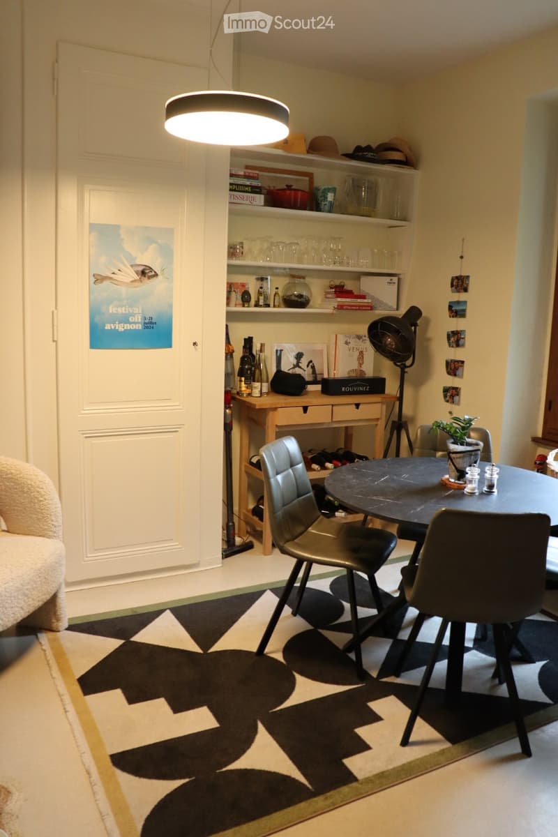Modern dining area with a round black table, four chairs, a shelf with glassware and books, a lamp, and a sofa in the background.