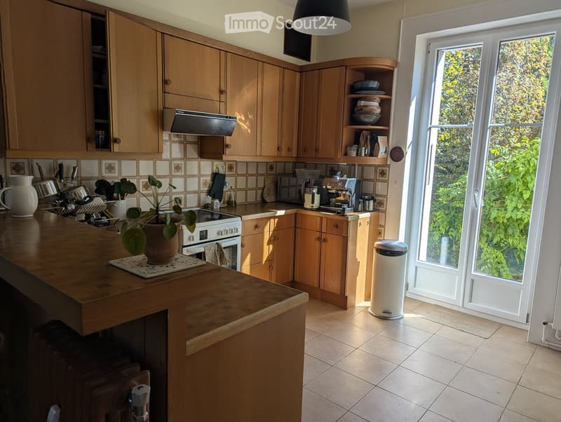 Bright kitchen with wooden cabinets, a central island, and a large window overlooking greenery. Appliances include an oven, coffee maker, and sink.