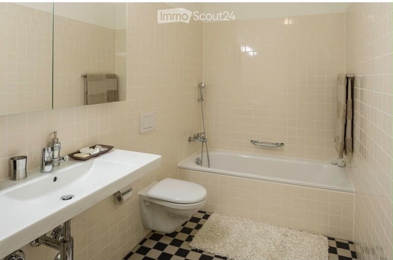 Bathroom with beige tiled walls, a white sink with a chrome faucet, a bathtub with a handheld shower, a toilet, a mirror, and a small white rug on the floor.