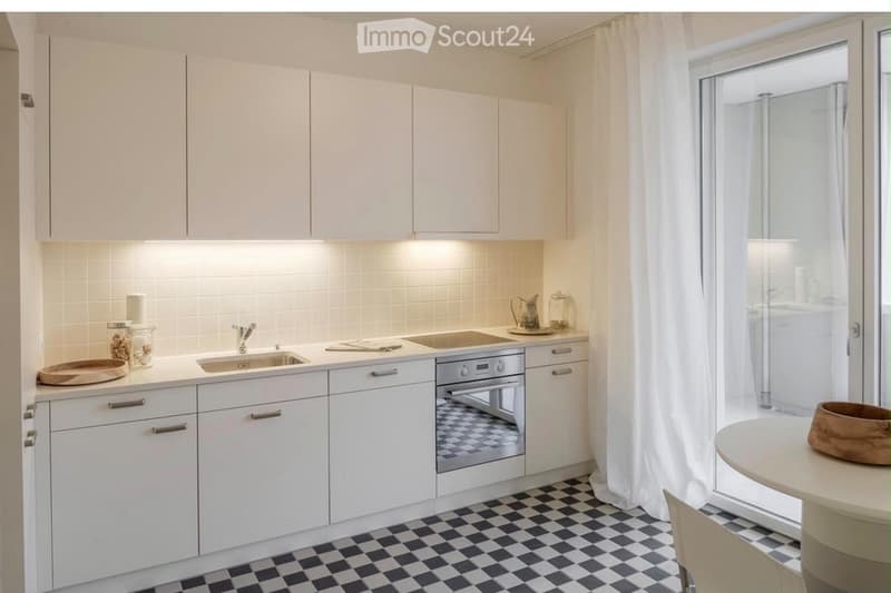 Modern kitchen with white cabinets, stainless steel sink, built-in oven, and a small table with a bowl in the adjacent space.