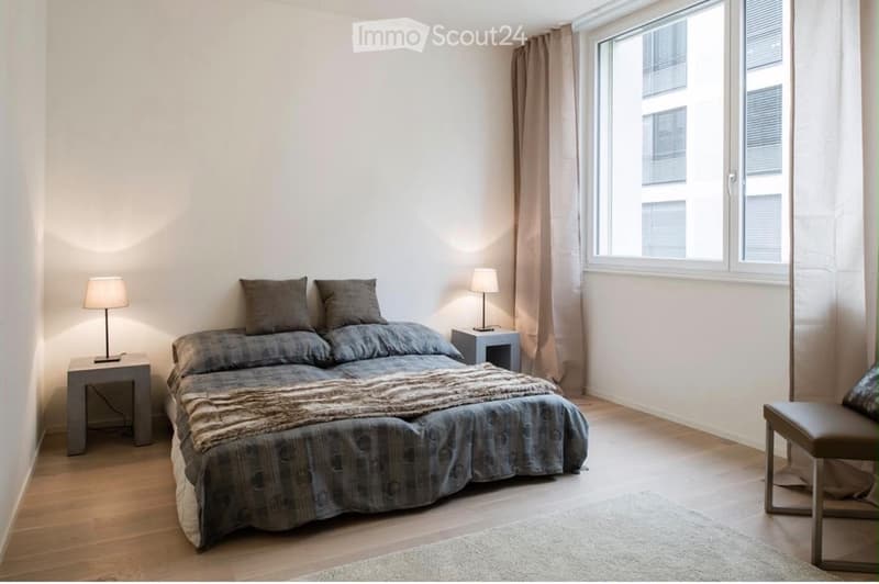 Modern bedroom with a large bed, nightstands, lamps, and a small stool near the window.