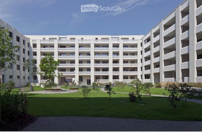 Modern multi-story residential building with white facade, multiple windows, and balconies. The courtyard features green lawns, trees, and benches.