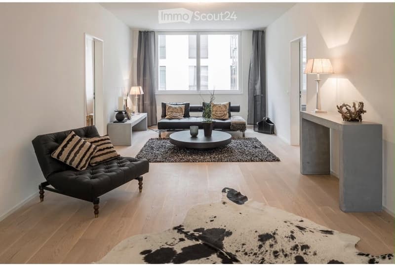 Modern living room with light wooden flooring, a black leather sofa, a round coffee table, a black leather chair, and decorative elements including a cowhide rug and table lamps.