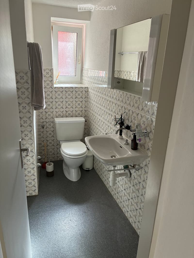 Small bathroom with white toilet, sink, and mirror. Decorative patterned tiles on walls, towel hanging on the side, and a window with frosted glass.