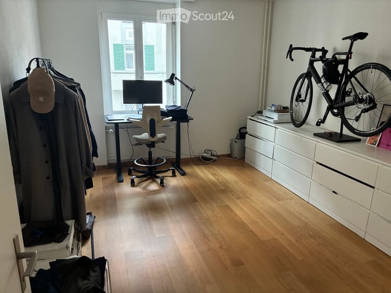 A modern, well-lit room with wooden flooring, a desk setup with a chair, a bicycle mounted on a stand, and a coat rack with a hat and coat.