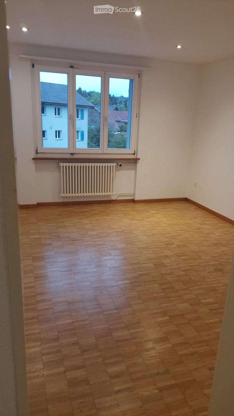 Empty room with wooden flooring, white walls, large windows, and a radiator below the windows.