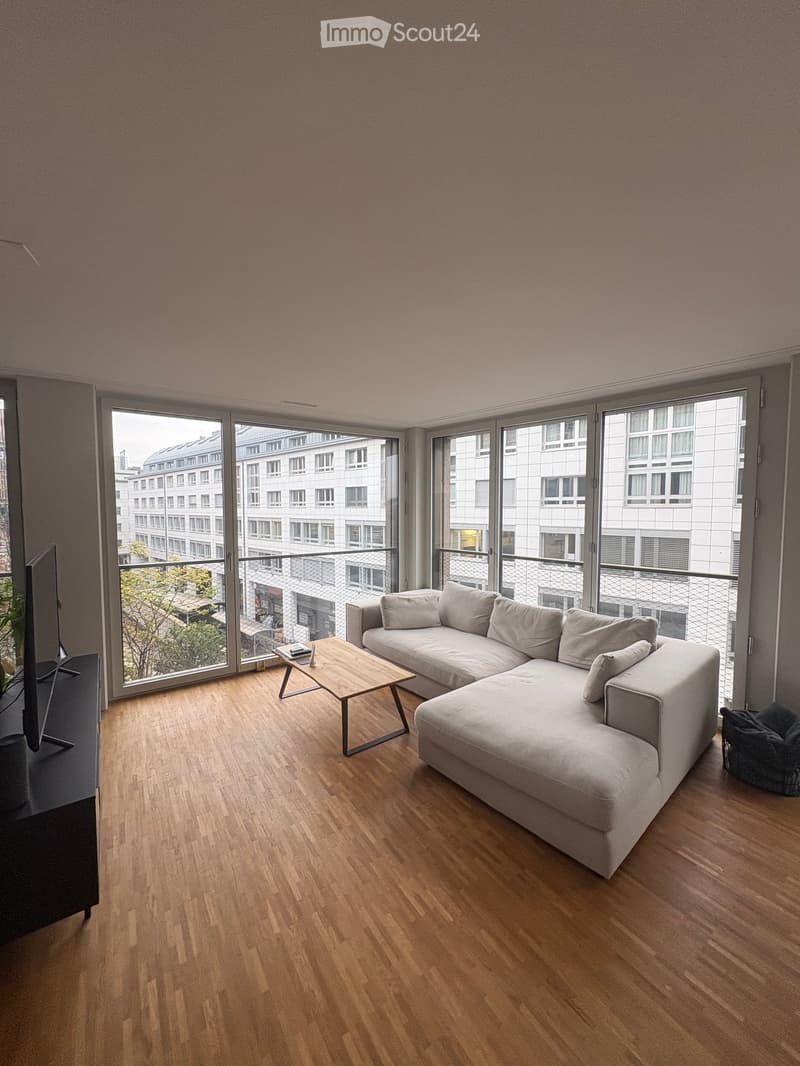 Modern living room with light wooden flooring, large windows, a grey sectional sofa, a wooden coffee table, and a TV stand.