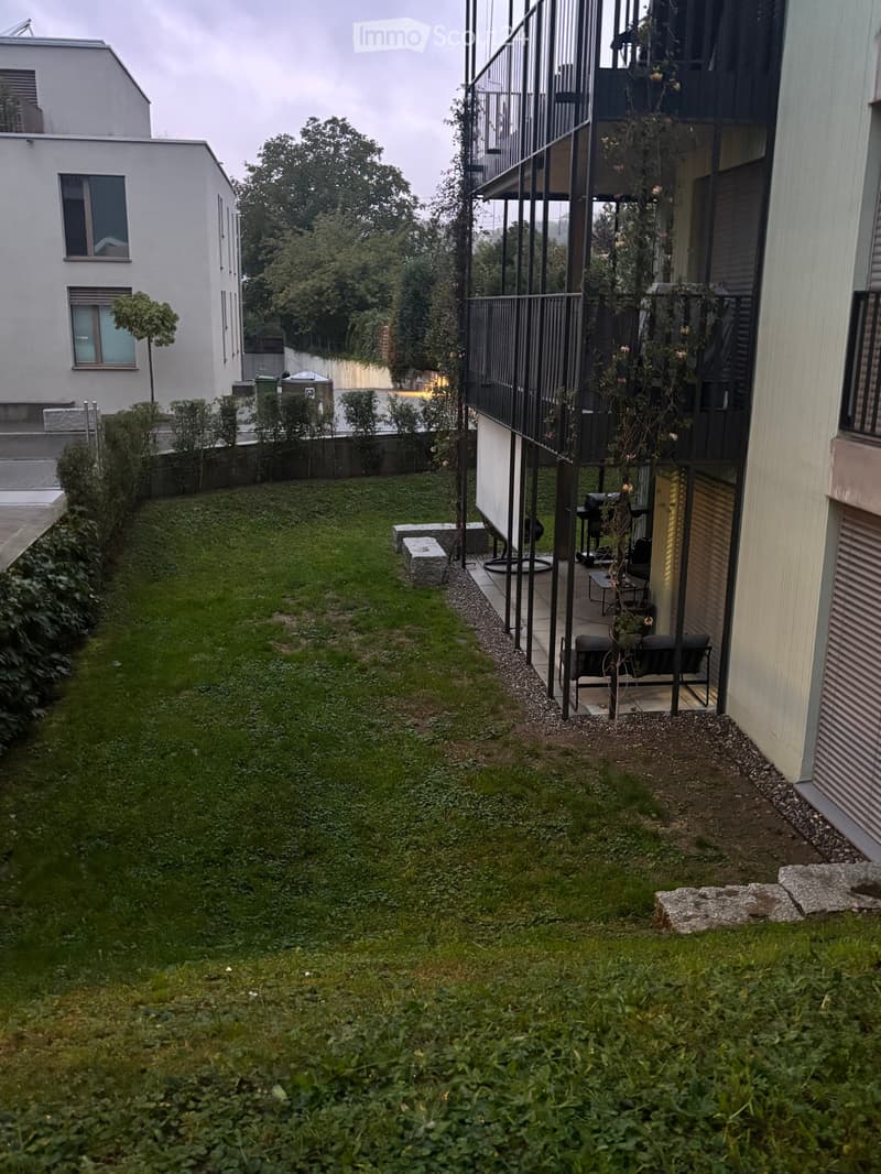 A narrow, grassy garden area with a modern balcony on the right side, featuring outdoor furniture and plants.