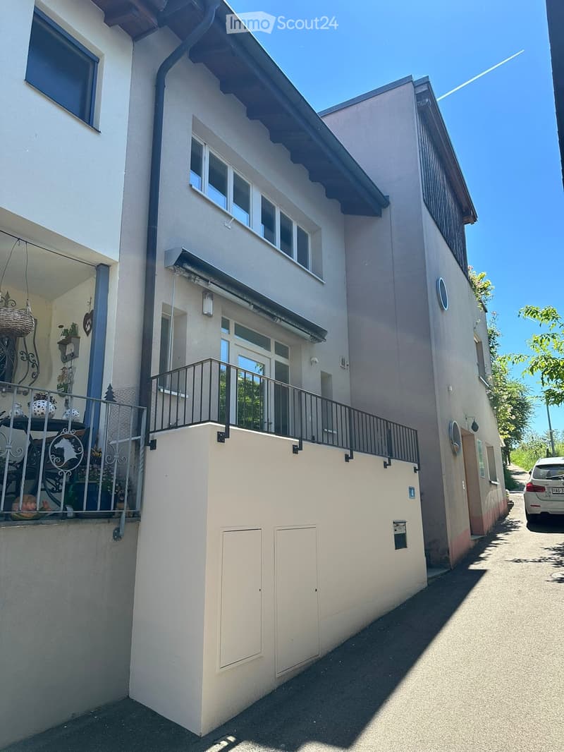 Two-story house with light beige exterior, multiple windows, and a balcony with metal railing. The house features a raised ground floor and a small garden area visible on the left side.