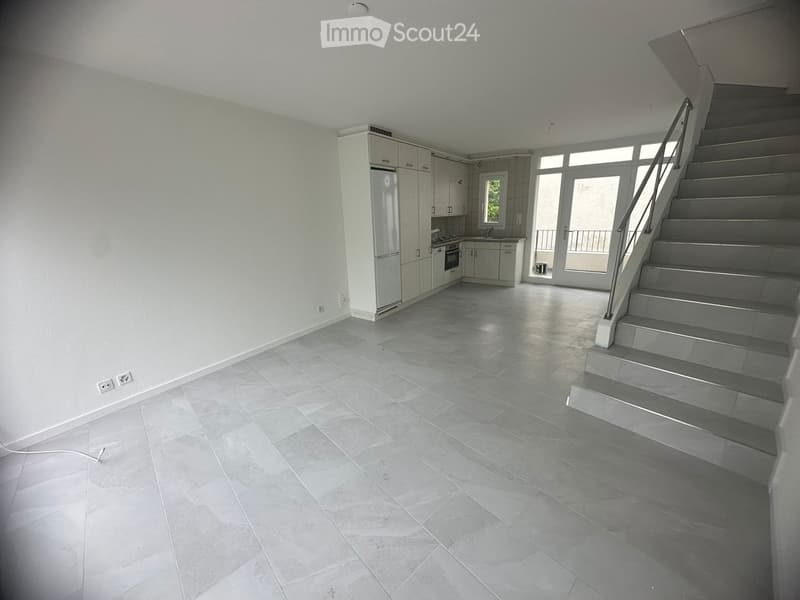 Empty room with grey tiled floor, white walls, staircase leading upwards, and a small kitchen area with cabinets and appliances.
