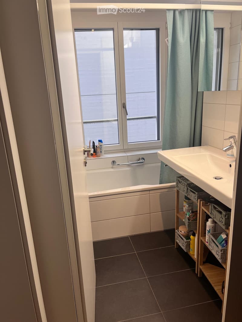 Bathroom with a bathtub, sink, and a small wooden shelf holding various items. The room has a window with a curtain and tiled flooring.