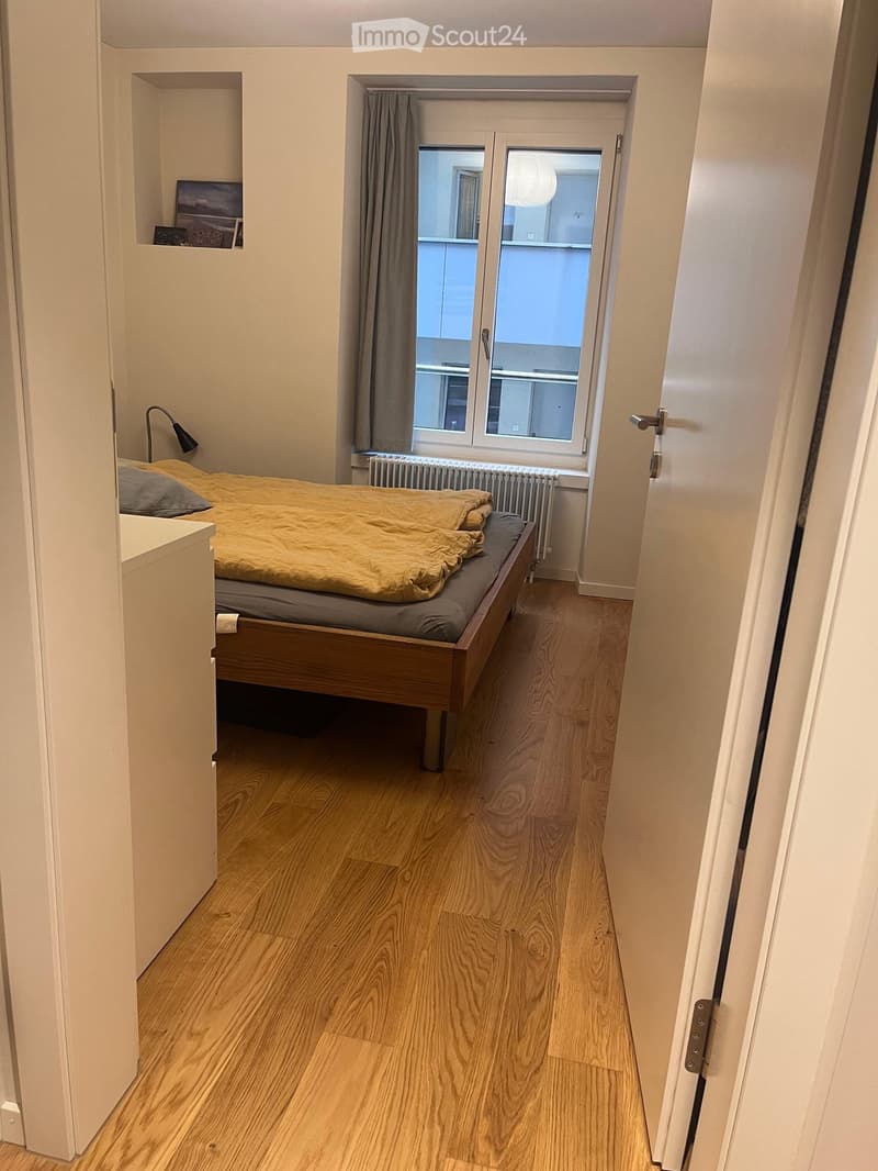Cozy bedroom with wooden floor, single bed with yellow bedding, nightstand, lamp, radiator, and window with curtain.