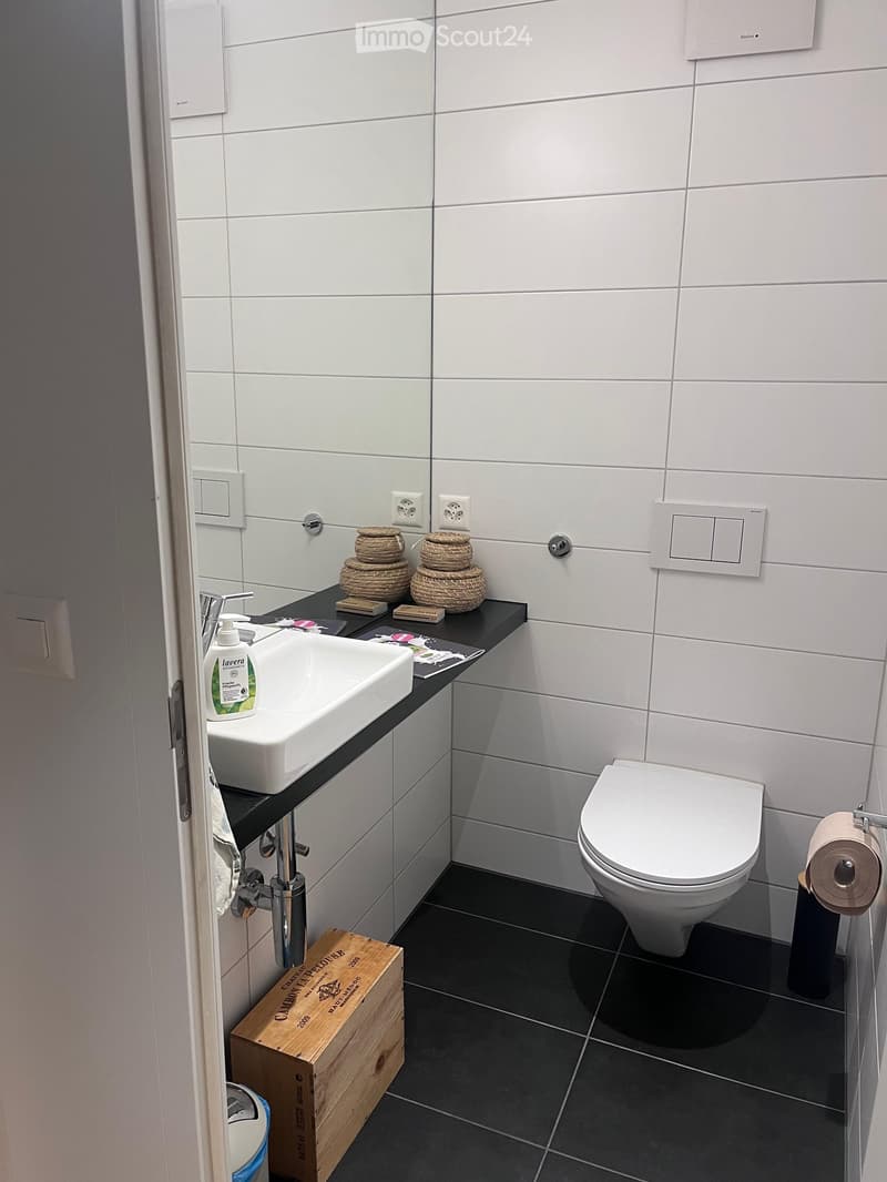 Modern bathroom with white tiles, black countertop, white sink, toilet, wooden crate, and decorative wicker baskets.