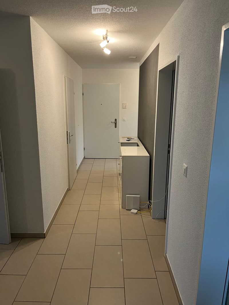 Empty hallway with tiled floor, white walls, and several doors. A small utility area with a sink and washing machine is visible on the right side.