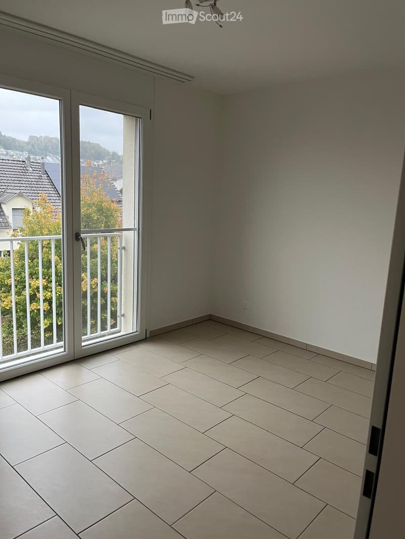 Empty room with tiled floor, white walls, large sliding glass doors leading to a balcony with a metal railing and a view of the neighborhood.