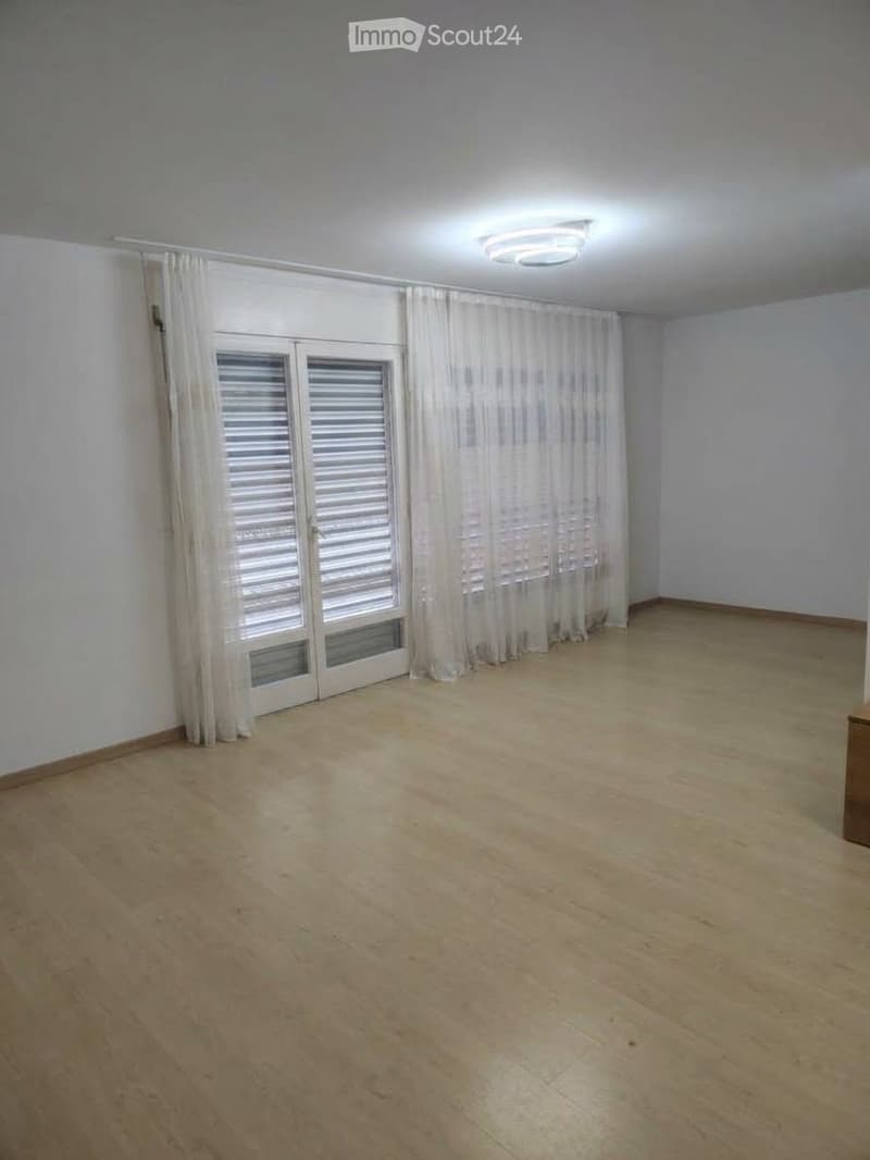 Empty room with wooden flooring, white walls, large window with white curtains, and a ceiling light fixture.