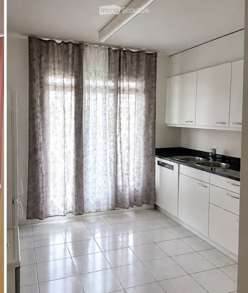 Modern kitchen with white cabinets, black countertops, and a stainless steel sink. The space features tiled flooring and large windows covered by patterned curtains.