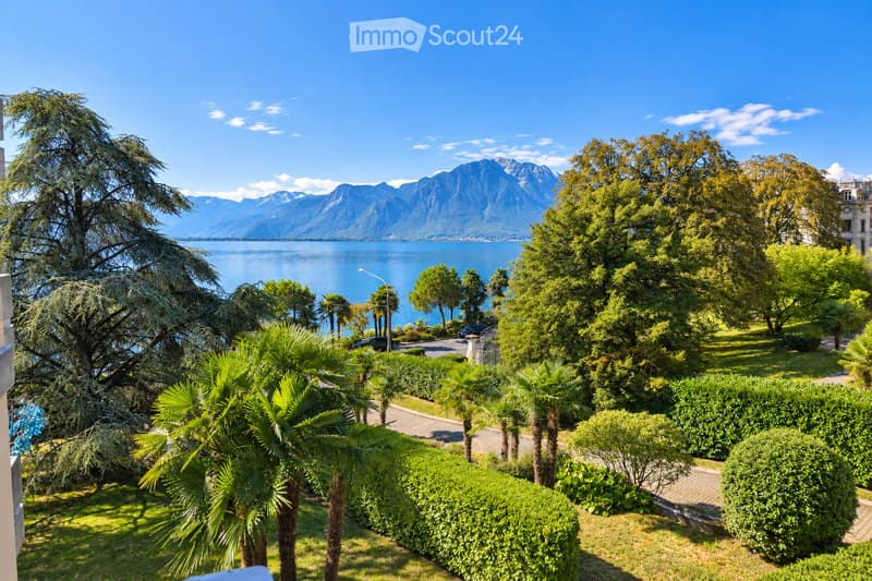 A lush garden with palm trees, neatly trimmed hedges, and a scenic view of a lake and mountains in the background.