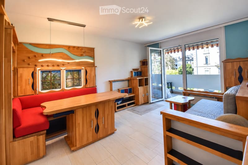 A modern, well-lit living space with a red sofa, wooden table, and a bed area, featuring built-in wooden cabinets and shelves.