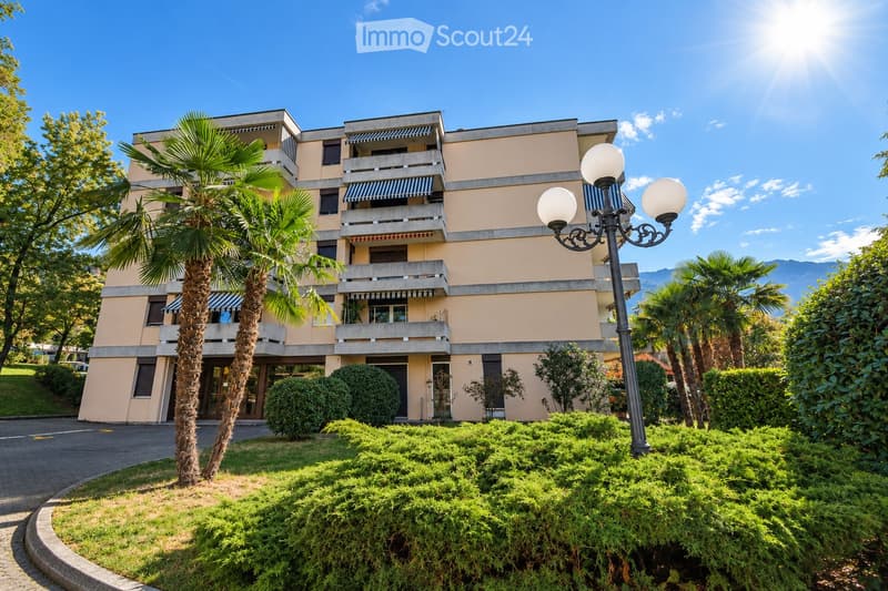 A multi-story residential building with beige walls, balconies, and striped awnings. The building is surrounded by well-maintained greenery, including palm trees and shrubs, with a decorative street lamp in the foreground.