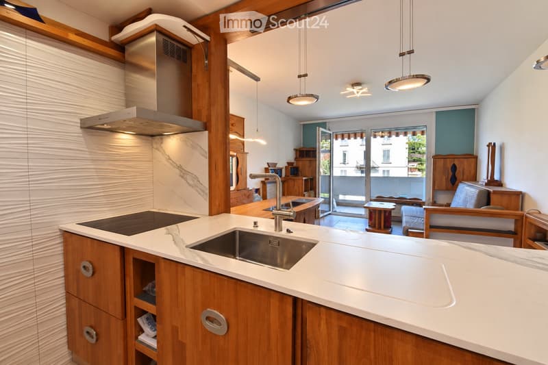 Modern kitchen with white marble countertops, wooden cabinets, stainless steel sink and faucet, built-in stove, and pendant lights. Features an open layout with a small seating area and a view to a balcony.