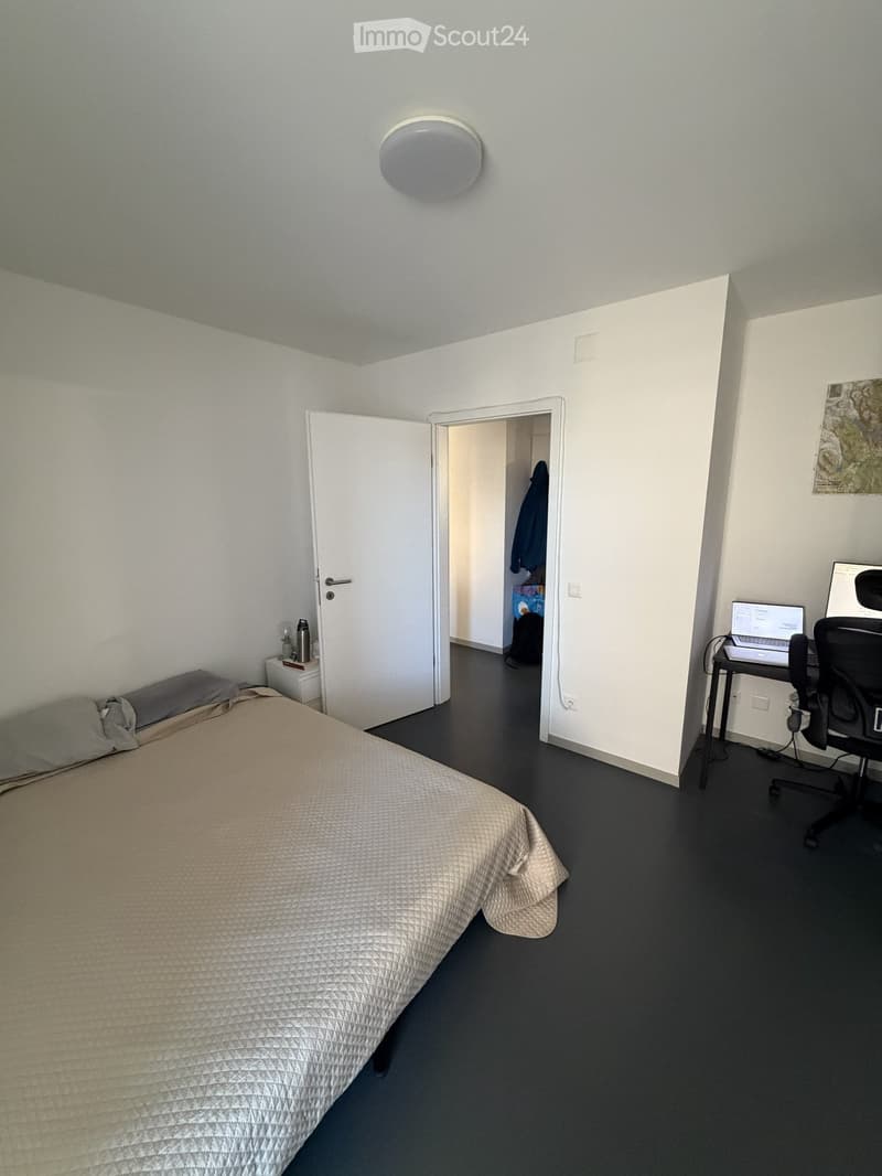 A modern bedroom with a neatly made bed, a desk with a laptop and chair, a nightstand with a lamp, and an open closet with clothes visible.