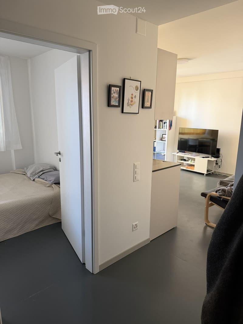 A modern, compact bedroom with a bed, a chair, a TV, a shelf, and picture frames on the wall.