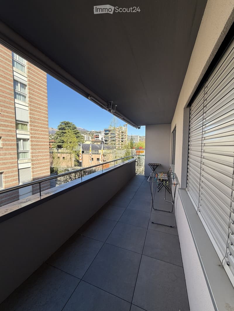 Balcony with tiled floor, metal railing, small table and chair, overlooking a cityscape with buildings and trees.