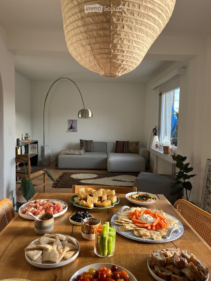 A cozy living room with a grey sofa, a wooden dining table set with various food items, a floor lamp, and a bookshelf in the background.