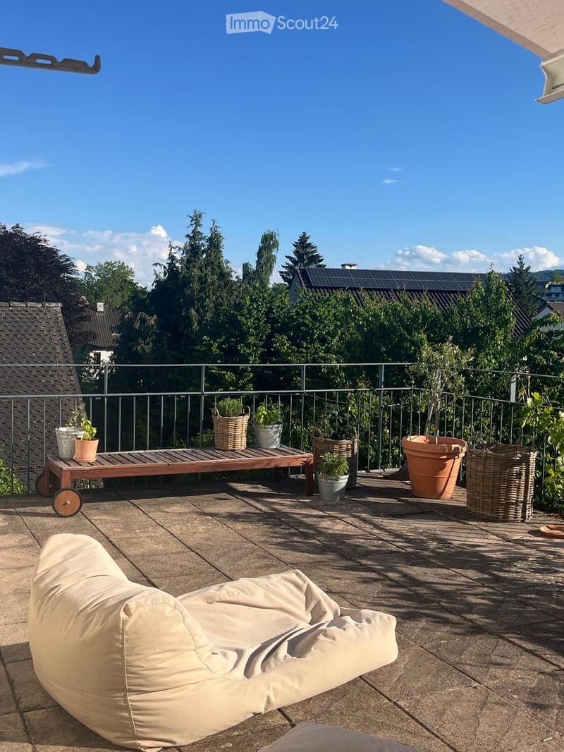 Balcony with a beige sofa, wooden table, potted plants, and a metal railing overlooking a green area.