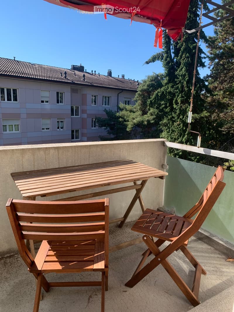 Balcony with wooden table and two wooden chairs, umbrella, overlooking a building and trees.