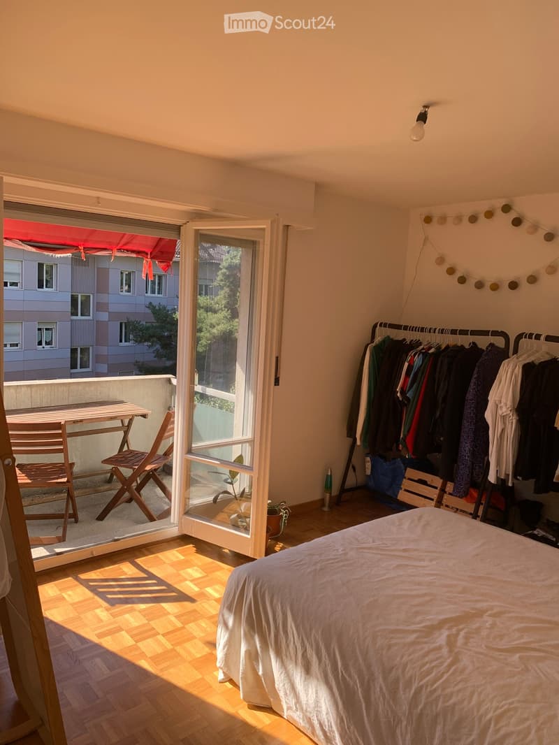Bright bedroom with wooden flooring, large window opening to a balcony, bed, wardrobe, and small table with chairs on the balcony.