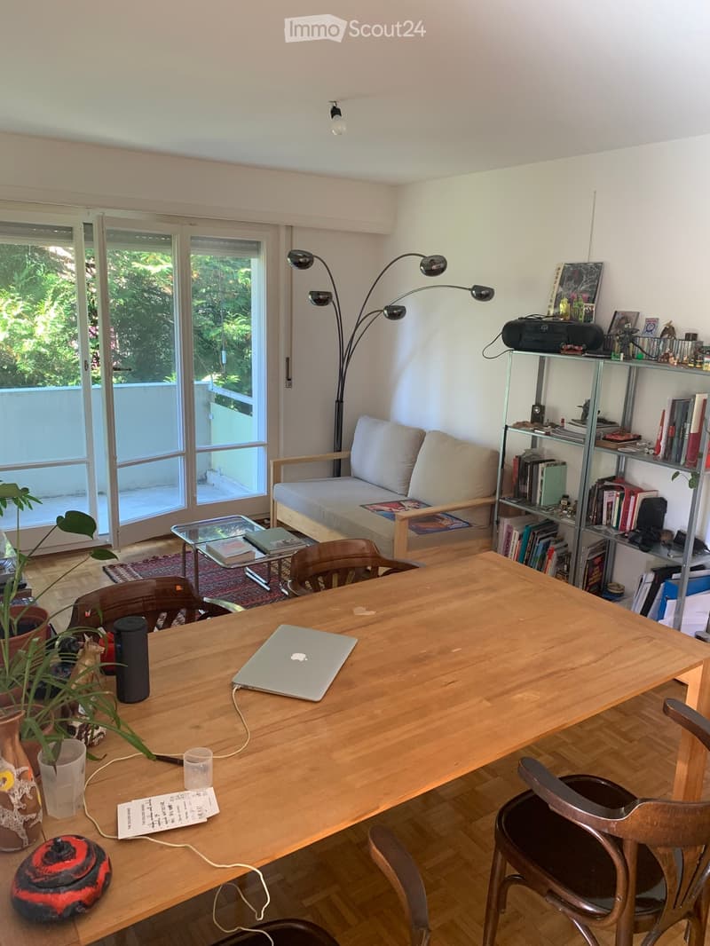 Bright living room with a wooden table, sofa, bookshelf, and large windows leading to a balcony.