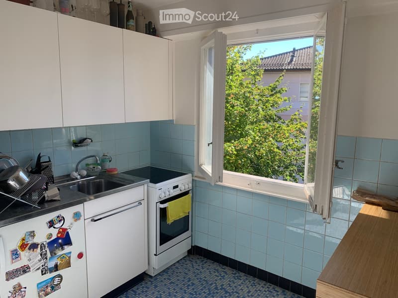 A compact kitchen with white cabinets, blue tiled backsplash, and a window overlooking greenery. Features include a refrigerator with various magnets, an oven with a yellow oven mitt, a sink with a faucet, and a dish rack with dishes.