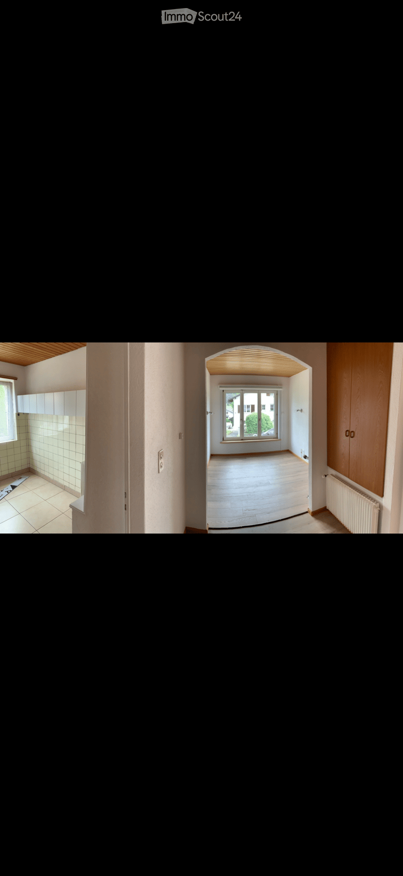 Empty hallway with wooden flooring, white walls, and a view into an adjacent room with large windows and a radiator.