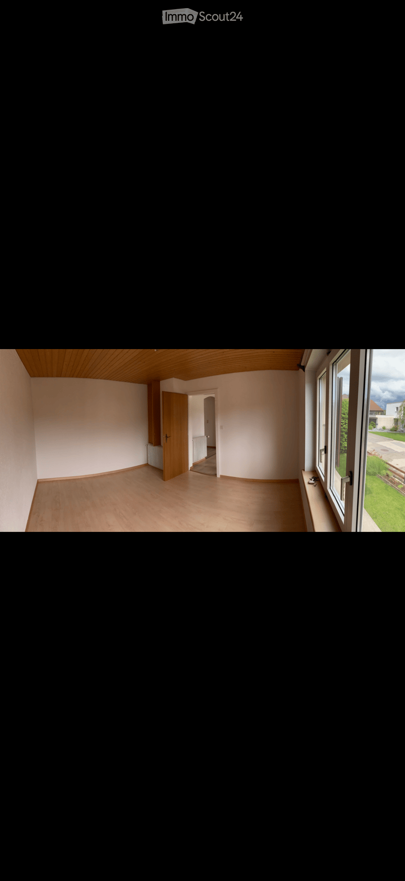 Empty room with wooden flooring, white walls, and large windows offering a view of a green garden. The room features a wooden door leading to another area and a slanted wooden ceiling.