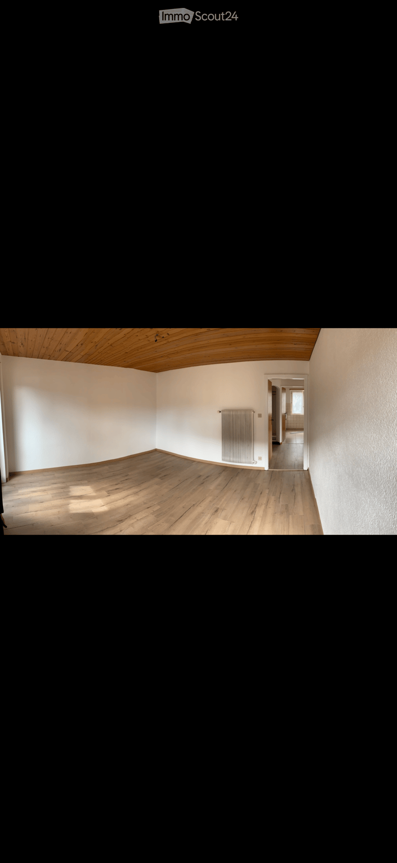 Empty room with wooden flooring, white walls, and a radiator. The room has a wooden slatted ceiling and a doorway leading to another room.