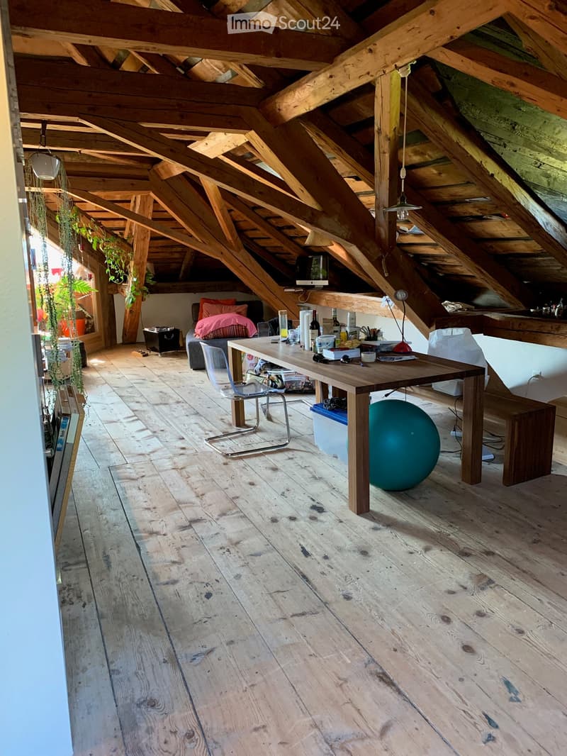 Loft-style room with exposed wooden beams and wooden flooring, featuring a long wooden table with chairs, a large exercise ball, and various items on the table including bottles and a lamp.