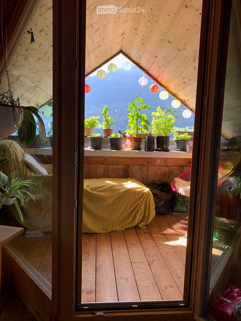 Cozy wooden balcony with a view of mountains, featuring a yellow sofa, small table, and several potted plants.